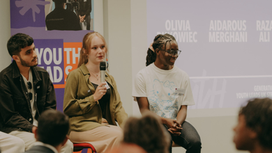 An image of three people sat on stools in front of a small crowd. The girl in the middle is holding a microphone and is speaking.