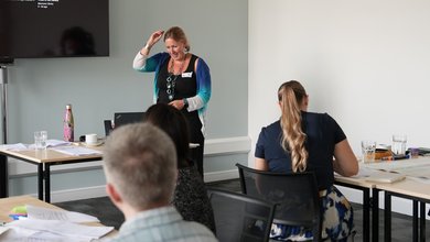 An image of someone delivering training to a group. There two other people sat at desks listening.